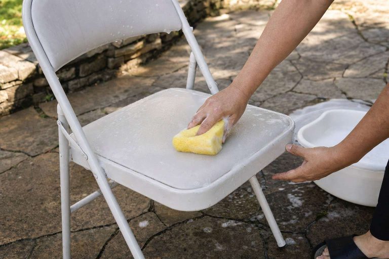 Hands scrubbing a white plastic folding chair clean with a soapy sponge on a patio