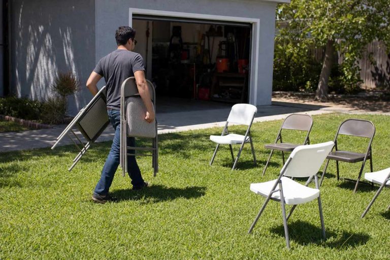A person carrying folding chairs across a lawn while white resin and gray metal folding chairs stand unfolded nearby